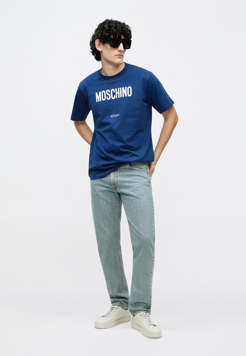 Blauw katoenen T-shirt met daarop de teksten "MOSCHINO" en "Milano" in het wit, gecombineerd met lichtblauwe spijkerbroek en witte sneakers. Model draagt een zonnebril.