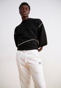 Sort sweater med en struktureret overflade og hvide accentlinjer, parret med hvide sweatpants med et logo på venstre ben.