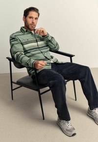 Man wearing green striped shirt, dark jeans, and gray sneakers sitting on black chair against plain beige background.