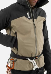 Veste imperméable noire et beige, avec un col haut, fermeture éclair avant et poches latérales. Comprend des boucles en métal et un harnais d'escalade.