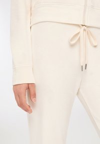 Osoba w jasnobeżowych spodniach z troczkiem związanym w kokardę oraz pasującą bluzką z długim rękawem, z ręką swobodnie opartą na boku.