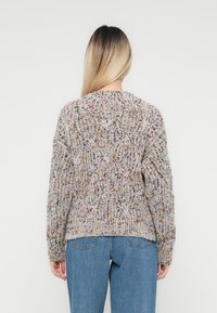 Vrouw met lang blond haar, gekleed in een meerkleurige gebreide trui en blauwe jeans, die met haar rug naar de camera staat tegen een effen witte achtergrond.