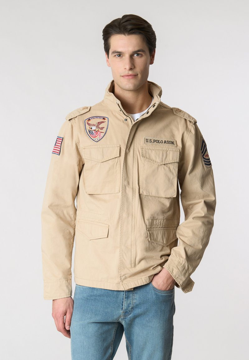 Giacca militare beige con colletto alto, diverse tasche frontali e spalline. Caratterizzata da patch ricamate e una bandiera americana.