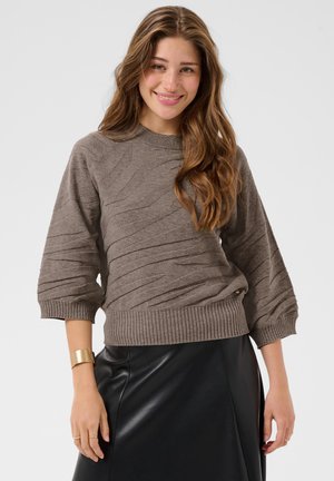 Jeune femme aux longs cheveux ondulés portant un pull marron texturé, une jupe en cuir noire et un bracelet manchette en or, souriante, regardant devant elle.