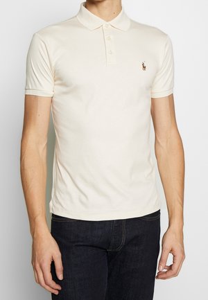 Homme portant un polo crème à manches courtes avec un petit logo brodé sur la poitrine gauche et un jean foncé.