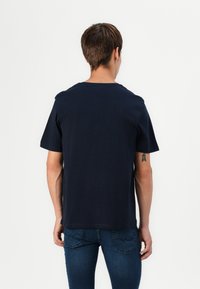 Camiseta de algodón azul marino de manga corta, con cuello redondo y corte recto, vista desde la espalda. Los detalles incluyen un tatuaje en el brazo izquierdo.