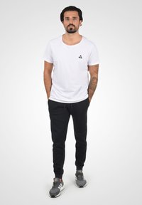 Hvid crew-neck t-shirt med et lille sort, trekantet logo på brystet, parret med sorte joggers og grå sneakers med hvide såler.