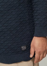 Navy gebreide trui met een textuur van een diamanten patroon, geribbelde mouwen en zoom, met een klein zwart label aan de onderrand.