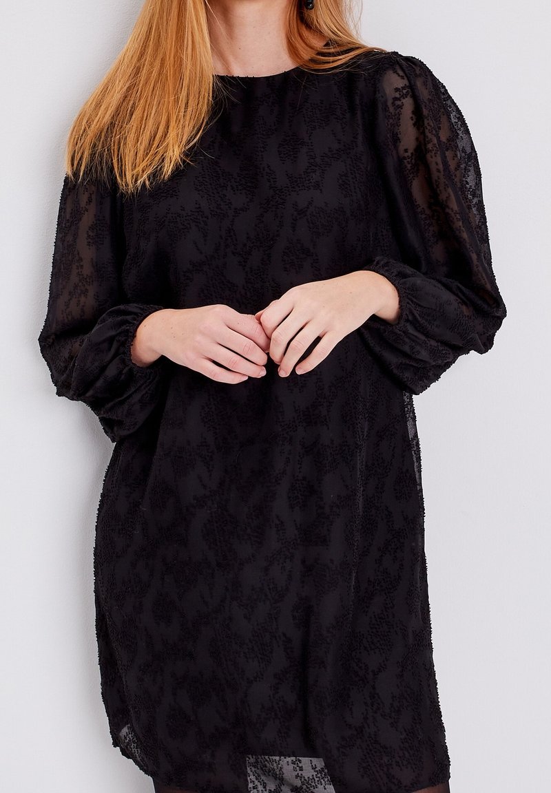 Black dress made of sheer fabric, featuring a textured floral pattern, puff sleeves, and a round neckline. Styled with a relaxed fit.