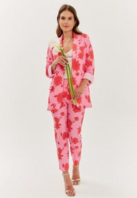 Jasně růžový oblek s červenými květinovými vzory, který zahrnuje strukturovanou blejzr a přiléhavé kalhoty. Modelka drží bílé květiny a má na sobě páskové lodičky.
