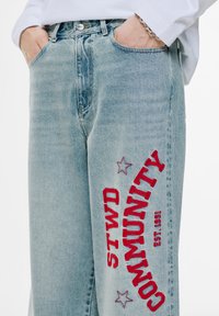 Ljusblå jeans i denim med lös passform, med röd brodyrtext "STWD COMMUNITY" och stjärnor på vänster ben.