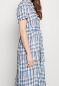 Žena v modro-bílé kostkované šatech s krátkým rukávem a odpovídajícím páskem, stojící bokem proti jednobarevnému pozadí.