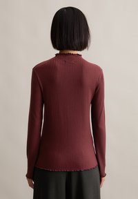 Langarm-Burgunderoberteil mit gerippter Textur, das einen Rüschenausschnitt und einen leicht abgerundeten Saum hat. Der Stoff wirkt weich und enganliegend.