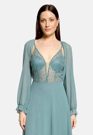 Femme portant une robe bleu sarcelle avec des manches longues transparentes et un corsage à décolleté plongeant en V orné de perles, debout devant un fond clair uni.