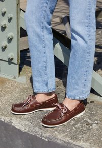 Scarpe da barca in pelle marrone con suole bianche e lacci rosa, cuciture dettagliate e occhielli dorati, indossate con jeans dritti blu chiaro.