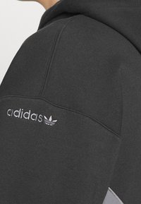 Närbild av svart Adidas-hoodie som visar vit Adidas-logotyp och broderad trefolddesign på axeln.