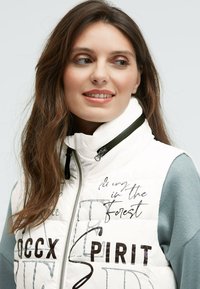 Weiße gepolsterte Weste mit hohem Kragen, die schwarze Schriftzüge und Grafiken zeigt. Graue Ärmel und ein Reißverschluss schließen die Vorderseite. Glatte Textur.