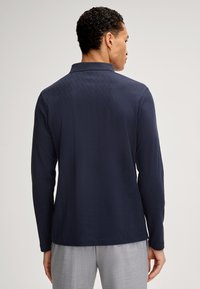 Camicia a maniche lunghe di colore blu navy con un motivo a diamante testurizzato, dotata di colletto piatto e orlo discreto, indossata con pantaloni grigi chiari.
