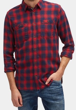 Mężczyzna w czerwono-granatowej koszuli w kratę z podwiniętymi rękawami i niebieskich dżinsach, z ręką w kieszeni.