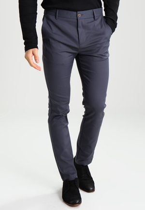 Hombre vistiendo pantalones ajustados azul marino y zapatos de vestir negros, de pie con una mano en el bolsillo contra un fondo liso.