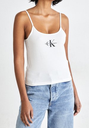 Kobieta ubrana w biały top na cienkich ramiączkach z czarnym logo "Calvin Klein Jeans" i jasnoniebieskie dżinsy na jednolitym tle.