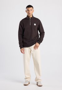 Brown zip-neck sweatshirt made of soft fabric, paired with light beige pants. Notable branding detail on the chest and neutral footwear.