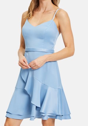 Femme portant une robe sans manches bleu clair avec des fines bretelles, un décolleté en V, une ceinture ajustée à la taille et une jupe à volants superposés.