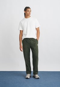 Witte T-shirt met korte mouwen en een relaxte fit, gecombineerd met donkergroene joggingsbroeken met twee zakken en een klein logo. Neutrale sneakers.