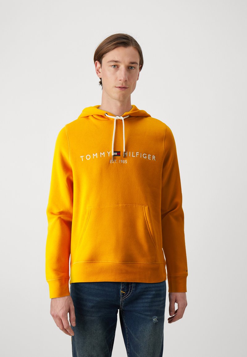 Žltá bavlnená mikina s kapucňou a sťahovacou šnúrkou, predná vrecko a vyšívané logo "TOMMY HILFIGER EST. 1985". V kombinácii s modrými džínsami.