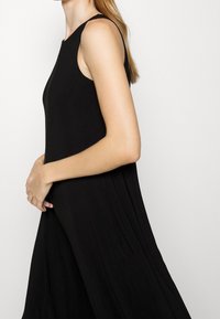 Svart ärmlös klänning med en lätt draperad silhuett och slät struktur. Har hög krage och minimal sömnad längs axeln.