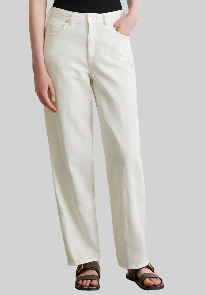 Białe, szerokie dżinsy wykonane z bawełny, z wysokim stanem, dwoma przednimi kieszeniami i zapięciem na guzik. Stylizowane z brązowymi sandałami.