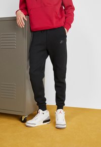 Zwarte Nike-sweatpants met een tailleband met trekkoord en zijzakken, gecombineerd met wit-grijze Air Jordan sneakers. Rode hoodie zichtbaar.
