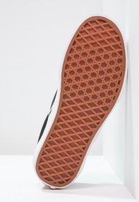 Semelle de chaussure en caoutchouc marron à motif gaufré, partie supérieure noire et blanche partiellement visible, positionnée verticalement contre des murs d’angle blancs.