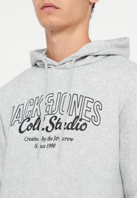 Sudadera con capucha gris que presenta un gran logo impreso en negro con el texto "JACK & JONES Col. Studio" y "Creado por el equipo Je desde 1990."