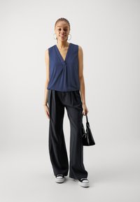 Top senza maniche blu navy con scollatura a V; abbinato a pantaloni neri a gamba larga e sneaker bianche; tiene in mano una piccola borsa nera.