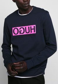 Man som bär en marinblå sweatshirt med en ljusrosa rektangulär logotyp och omvänd texten "HUGO", lager på lager över en vit skjorta.