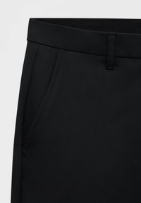Détail d'un pantalon noir sur mesure montrant la taille, les passants de ceinture et la poche latérale avec des coutures visibles sur un fond uni.