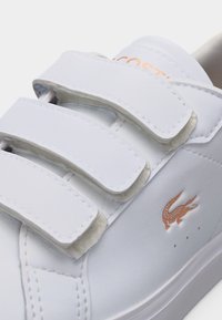 Λευκό αθλητικό παπούτσι με δύο λουριά βέλκρο και ένα μικρό πορτοκαλί λογότυπο κροκοδείλου στο πλάι κοντά στην σόλα.