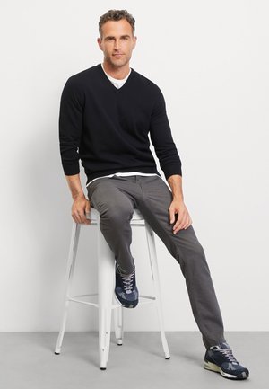 Pull noir à col en V porté sur une chemise blanche, pantalon gris et baskets bleu marine. Assis sur un tabouret blanc, coiffure courte et ondulée.