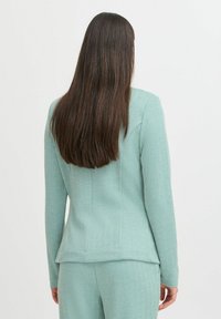 Mintgroene op maat gemaakte blazer met een gestructureerd visgraatpatroon. Lange mouwen en een getailleerd ontwerp dat versmalt bij de taille.