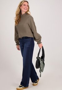 Vrouw die loopt met een zwarte handtas met een groene sjaal, in een bruin langemouwen shirt, donkerblauwe jeans en beige sneakers.