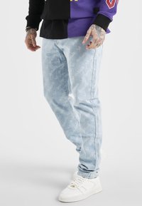 Ljusblå jeans med vitt textmönster, bärs med vita sneakers och en långärmad tröja som är hälften svart och hälften lila.