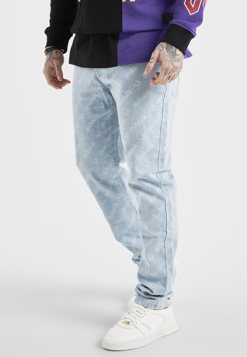 Ljusblå jeans med vitt textmönster, bärs med vita sneakers och en långärmad tröja som är hälften svart och hälften lila.