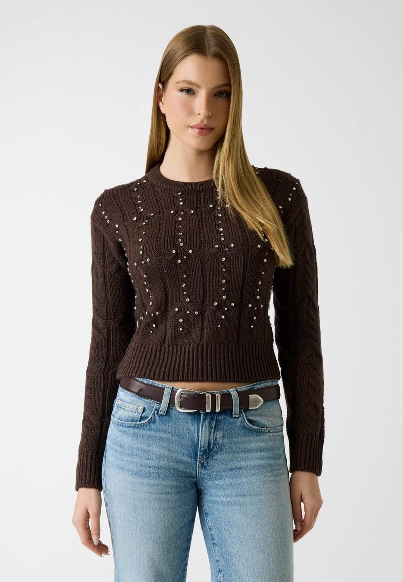 Femme aux longs cheveux blonds portant un pull en maille marron foncé orné de perles et un jean bleu clair avec une ceinture marron.