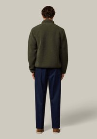 Vihreä fleecetakki, jossa korkea kaulus, yhdistettynä sinisiin housuihin ja ruskeisiin kenkiin, esitetty takaa neutraalia taustaa vasten.