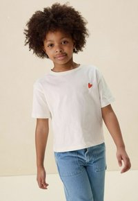 Witte T-shirt met korte mouwen, voorzien van een klein rood hartje op de linkerkant van de borst, gecombineerd met lichtblauwe spijkerbroek. Casual pasvorm en zachte stof.