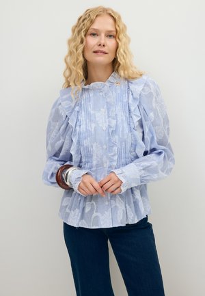 Vrouw met krullend blond haar draagt een blauw gestreept blouse met ruches en bloemenpatronen, gecombineerd met donkerblauwe jeans en grote armbanden aan één pols.