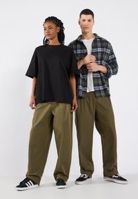 T-shirt noir surdimensionné, pantalon large vert olive, chemise à carreaux bleue et verte, et baskets noires avec des rayures blanches. Ensemble décontracté.