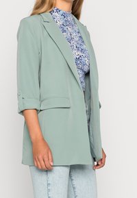 Miętowa marynarka z podwiniętymi rękawami, wykonana z gładkiego materiału. Posiada przednie kieszenie i dopasowany fason, noszona na kwiecistej bluzce.