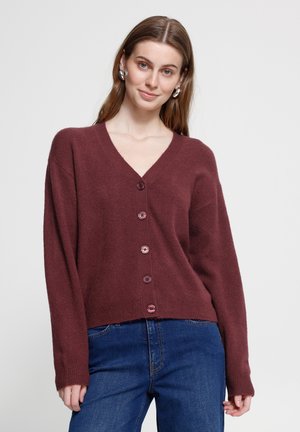 Femme aux longs cheveux bruns portant des boucles d’oreilles argentées en anneaux, un cardigan bordeaux boutonné et un jean bleu, debout devant un fond clair uni.